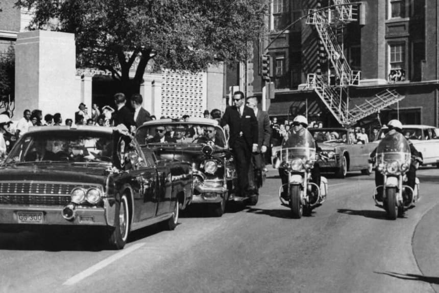 Vista a través del parabrisas del descapotable en primer plano, la mano del presidente John F. Kennedy se dirige hacia su cabeza pocos segundos después de recibir un disparo mortal, mientras la primera dama Jacqueline Kennedy le sujeta el antebrazo mientras la caravana avanza por Elm Street pasando por el Texas School Book Depository ,en Dallas, el 22 de noviembre de 1963. (James W. «Ike» Altgens/Foto AP)
