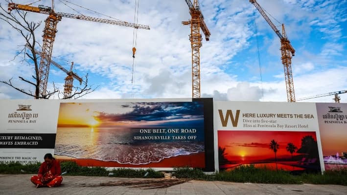 Una mujer sentada junto a una valla publicitaria de la Iniciativa de la Franja y la Ruta de China en Sihanoukville, Camboya, el 1 de julio de 2024. Disfrazada de desarrollo de infraestructuras, la plataforma de un billón de dólares se apodera de los recursos naturales de otros países y amplía el uso de sus puertos con fines civiles y militares. (Valeria Mongelli/Getty Images)