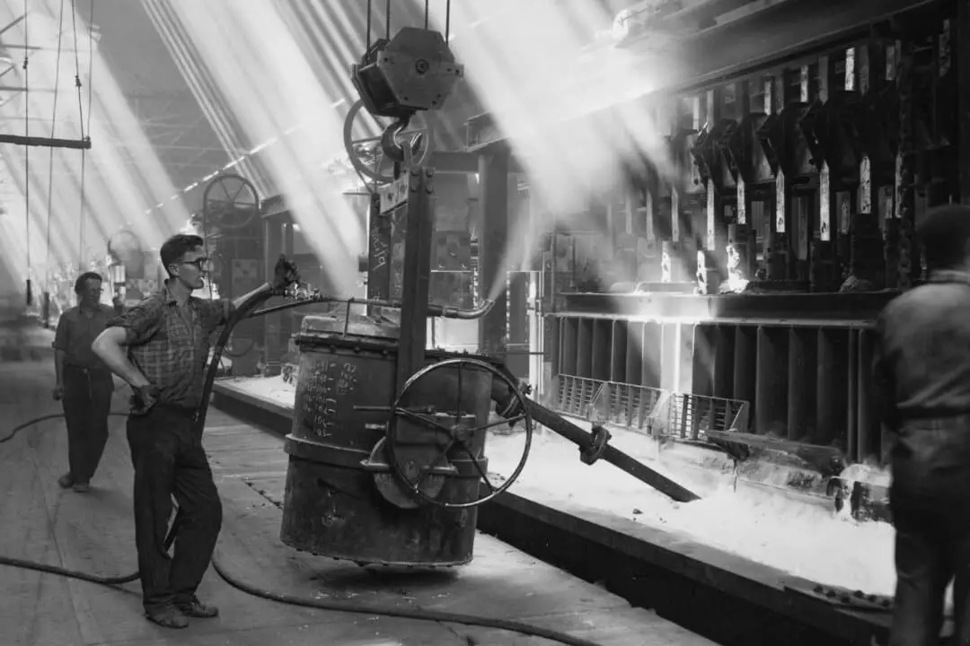 Un trabajador de una fábrica maneja una gran máquina suspendida de una polea en una planta industrial en medio de haces de luz, alrededor de 1950. (FPG/Archive Photos/Getty Images)