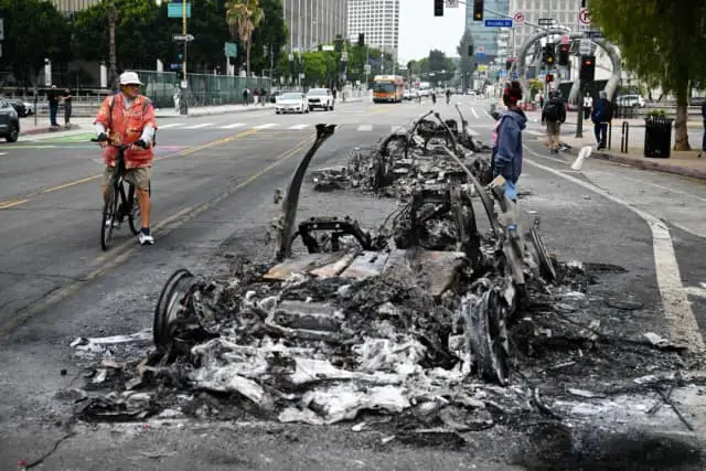 La gente observa los restos carbonizados de un vehículo quemado en la calle tras una noche de protestas en respuesta a las operaciones federales de inmigración en Los Ángeles, el 9 de junio de 2025. (Ronaldo Schemidt/AFP vía Getty Images).