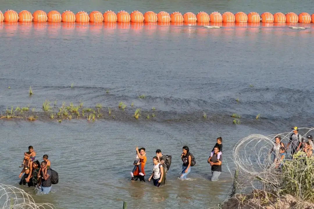 Personas caminan entre alambre de púas y una serie de boyas colocadas en el agua a lo largo de la frontera con México en Eagle Pass, Texas, el 16 de julio de 2023. (Suzanne Cordeiro/AFP/Getty Images)
