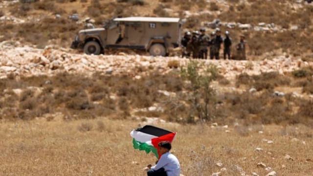 Un niño sostiene una bandera palestina mientras las fuerzas de seguridad israelíes montan guardia, en Raba, cerca de Jenin, en Cisjordania, el 18 de julio de 2025. (Raneen Sawafta/Reuters)
