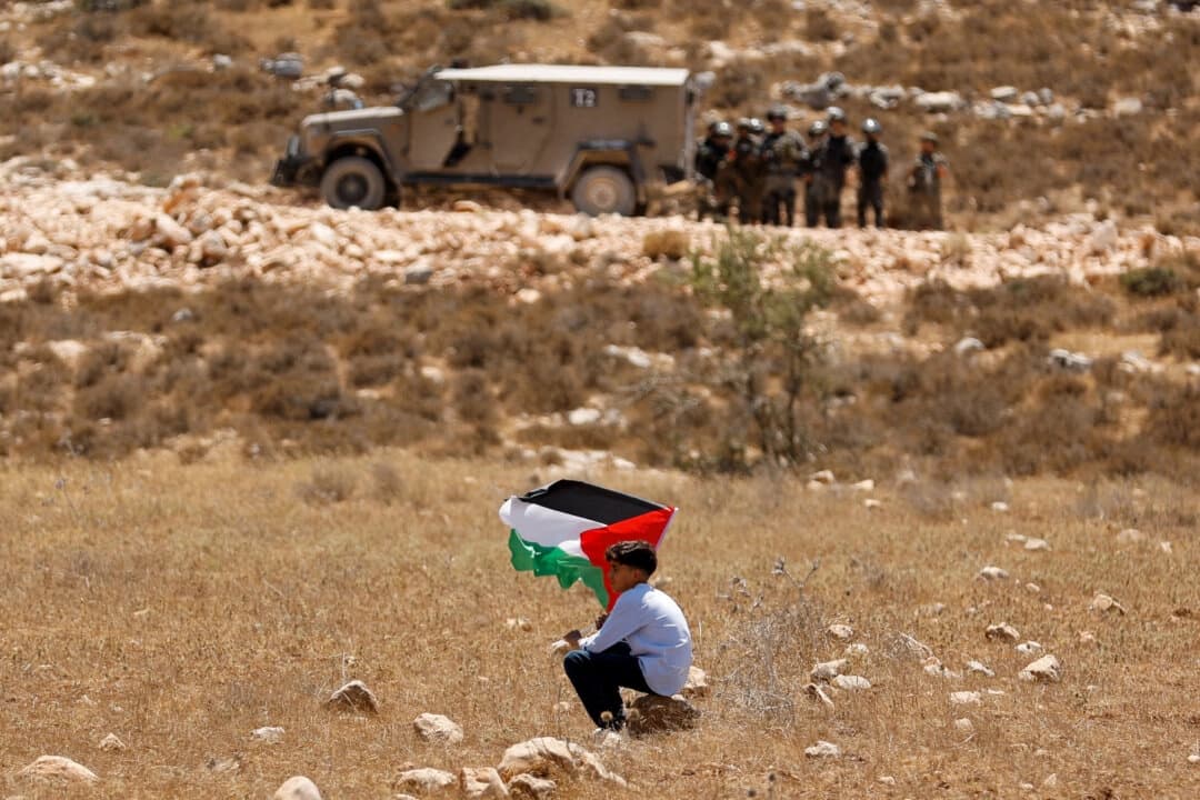 Un niño sostiene una bandera palestina mientras las fuerzas de seguridad israelíes montan guardia, en Raba, cerca de Jenin, en Cisjordania, el 18 de julio de 2025. (Raneen Sawafta/Reuters)