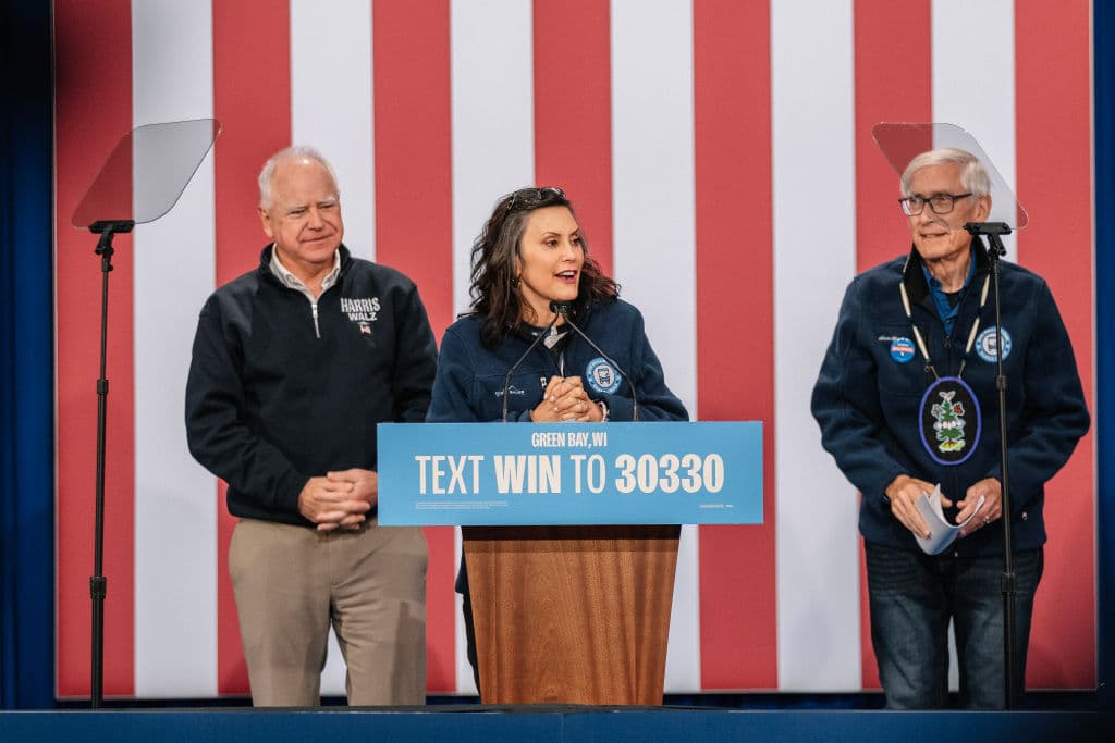 El candidato demócrata a la vicepresidencia, el gobernador de Minnesota Tim Walz (izquierda), y el gobernador de Wisconsin Tony Evers (derecha), escuchan a la gobernadora de Michigan Gretchen Whitmer (centro) hablar en un mitin para dar inicio a su gira en autobús «Driving Forward» Blue Wall Bus Tour en el centro de convenciones KI el 14 de octubre de 2024 en Green Bay, Wisconsin. (Foto de Jim Vondruska/Getty Images)