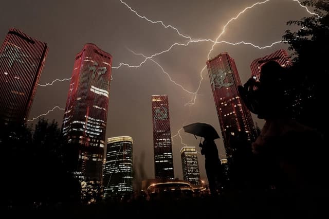Un rayo atraviesa el cielo mientras las personas contemplan los edificios iluminados con un espectáculo de luces en la víspera del centenario del Partido Comunista Chino en Beijing, el 30 de junio de 2021. (Noel Celis/AFP a través de Getty Images)
