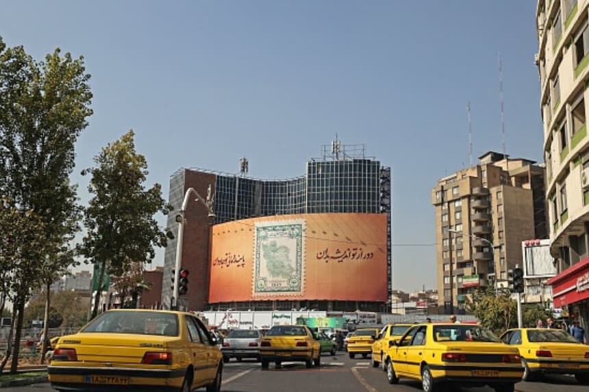 Taxis circulan por la plaza Valiasr, en el corazón de la capital iraní, Teherán, el 11 de octubre de 2022, pasando junto a una valla publicitaria con un sello postal que muestra el mapa de la República Islámica rodeado por la letra de una canción nacional. (ATTA KENARE/AFP a través de Getty Images).