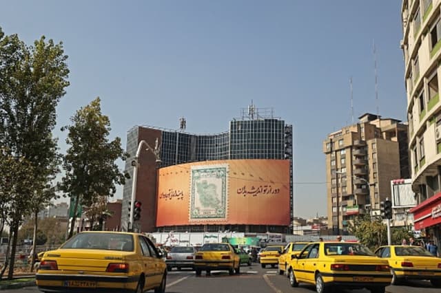 Taxis circulan por la plaza Valiasr, en el corazón de la capital iraní, Teherán, el 11 de octubre de 2022, pasando junto a una valla publicitaria con un sello postal que muestra el mapa de la República Islámica rodeado por la letra de una canción nacional. (ATTA KENARE/AFP a través de Getty Images).