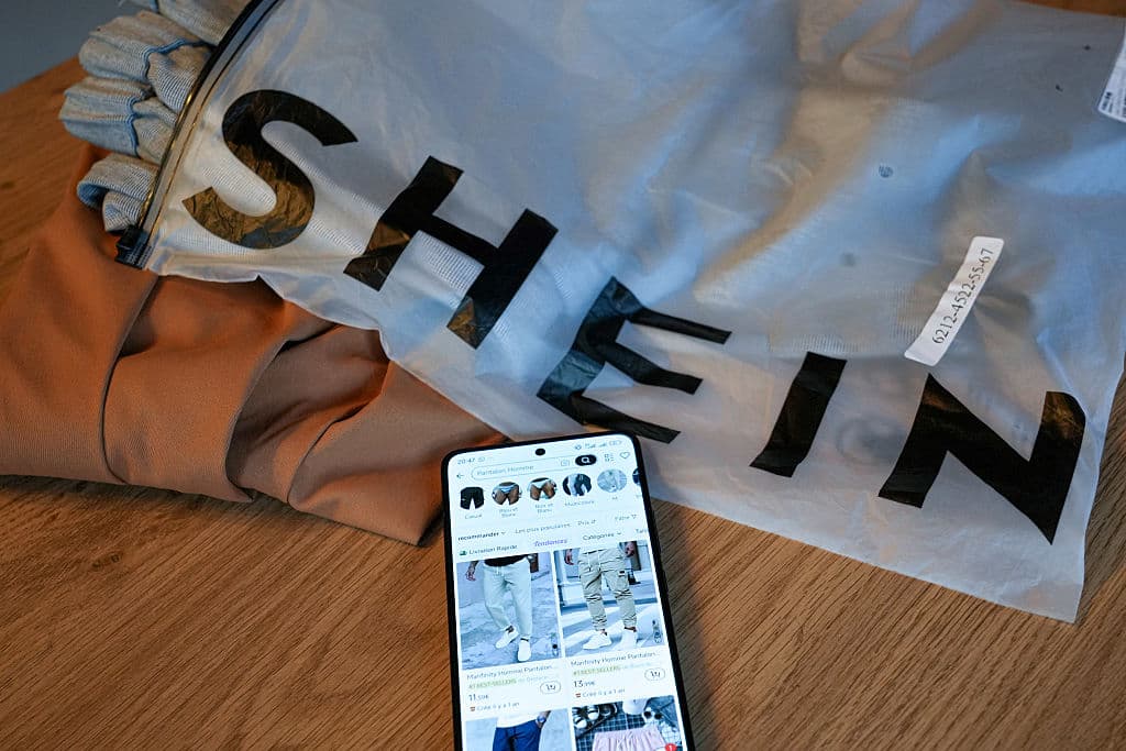 Un teléfono muestra artículos rebajados en la aplicación de venta de ropa online SHEIN sobre una bolsa de plástico de la marca china SHEIN que contiene una prenda de vestir sobre una mesa junto a pantalones de poliéster de baja calidad en Valence, Francia, el 3 de junio de 2025. (Foto de NICOLAS GUYONNET/Hans Lucas/AFP vía Getty Images)