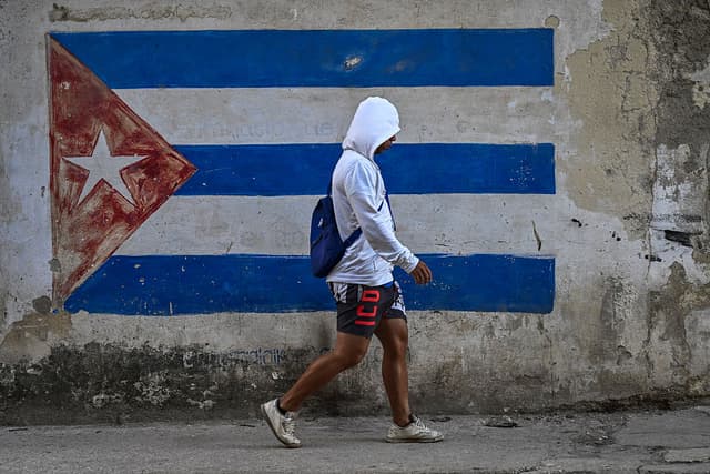 Un hombre pasa junto a un muro pintado con la bandera cubana en una calle de La Habana durante un apagón nacional el 22 de marzo de 2026. El Ministerio de Energía informó que el apagón afectó a toda la isla de Cuba el 21 de marzo, siendo el segundo apagón nacional en menos de una semana, mientras la red eléctrica sufre las consecuencias del bloqueo petrolero estadounidense. (Foto de YAMIL LAGE / AFP vía Getty Images)