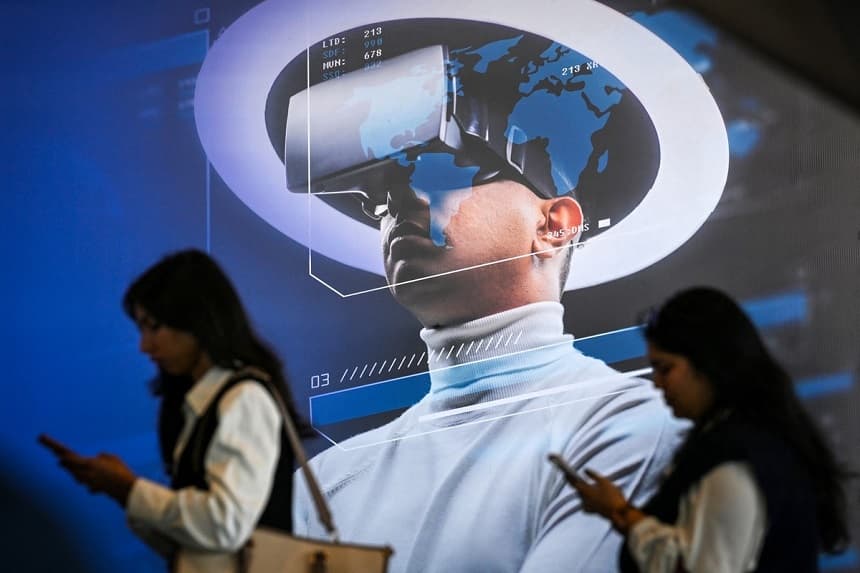 Los visitantes utilizan sus teléfonos móviles mientras pasan junto a una valla publicitaria que promociona unas gafas de realidad virtual en la Cumbre sobre el Impacto de la IA celebrada en Nueva Delhi, India, el 17 de febrero de 2026. (Arun Sankar/AFP vía Getty Images)
