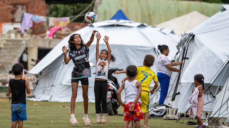 <em>Niños juegan con una pelota cerca de tiendas de campaña en el estadio de béisbol César Nieves en el barrio La Angustia de Catia La Mar, estado de La Guaira, Venezuela, el 3 de julio de 2026, donde se han refugiado personas desplazadas.  (Miguel MEDINA/AFP vía Getty Images)</em>