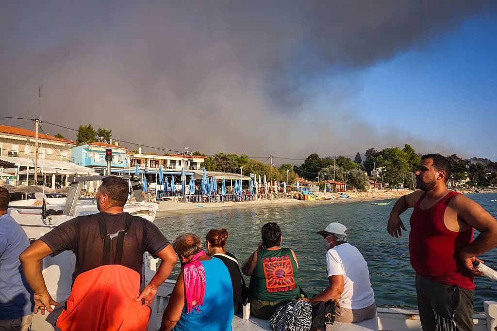 <em>Personas evacuan en pequeñas embarcaciones durante un incendio forestal mientras una columna de humo se eleva hacia el cielo cerca del pueblo de Siviri, en el norte de Grecia, el 13 de agosto de 2026. Una flota de pequeñas embarcaciones evacuó a cientos de turistas de las playas del norte de Grecia mientras un incendio forestal se acercaba a dos populares centros turísticos. (Vasilis Ververidis/AFP vía Getty Images).</em>