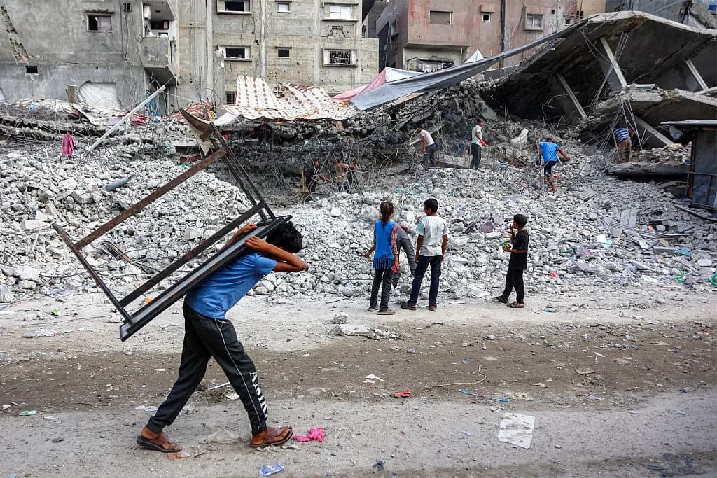 Un niño camina con una mesa a la espalda junto a trabajadores de recuperación que desmantelan los escombros de un edificio destruido durante la guerra de Israel contra Hamás en Gaza, con el fin de reutilizar los restos reciclados en el futuro, en la ciudad de Gaza el 16 de agosto de 2026. (Omar Al-Qattaa/AFP vía Getty Images).