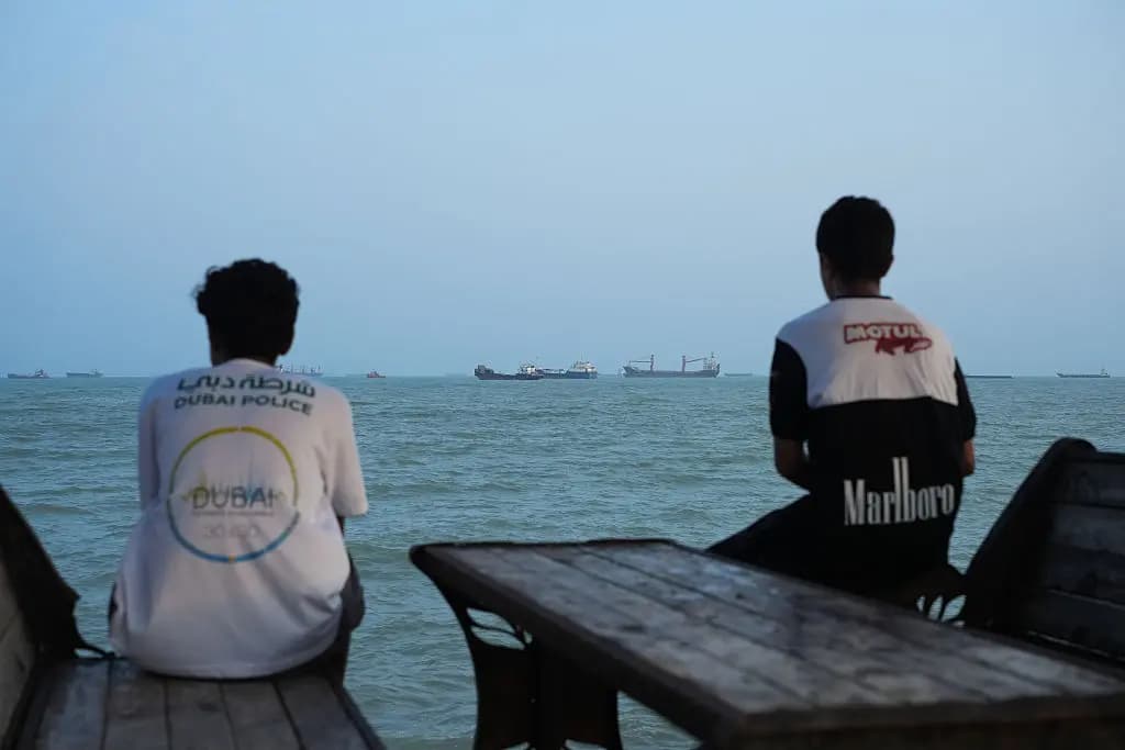Dos jóvenes están sentados en la orilla mientras se ven barcos anclados en el Estrecho de Ormuz, frente a la costa de Bandar Abbas, Irán, el 10 de agosto de 2026. (Ali Saeedi/Getty Images).