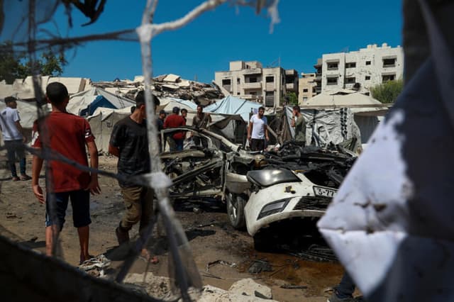 Residentes caminan por una zona dañada junto a un coche destruido tras los ataques israelíes en la ciudad de Gaza el 1 de septiembre de 2026. (Eyad Azam/Middle East Images/AFP vía Getty Images).