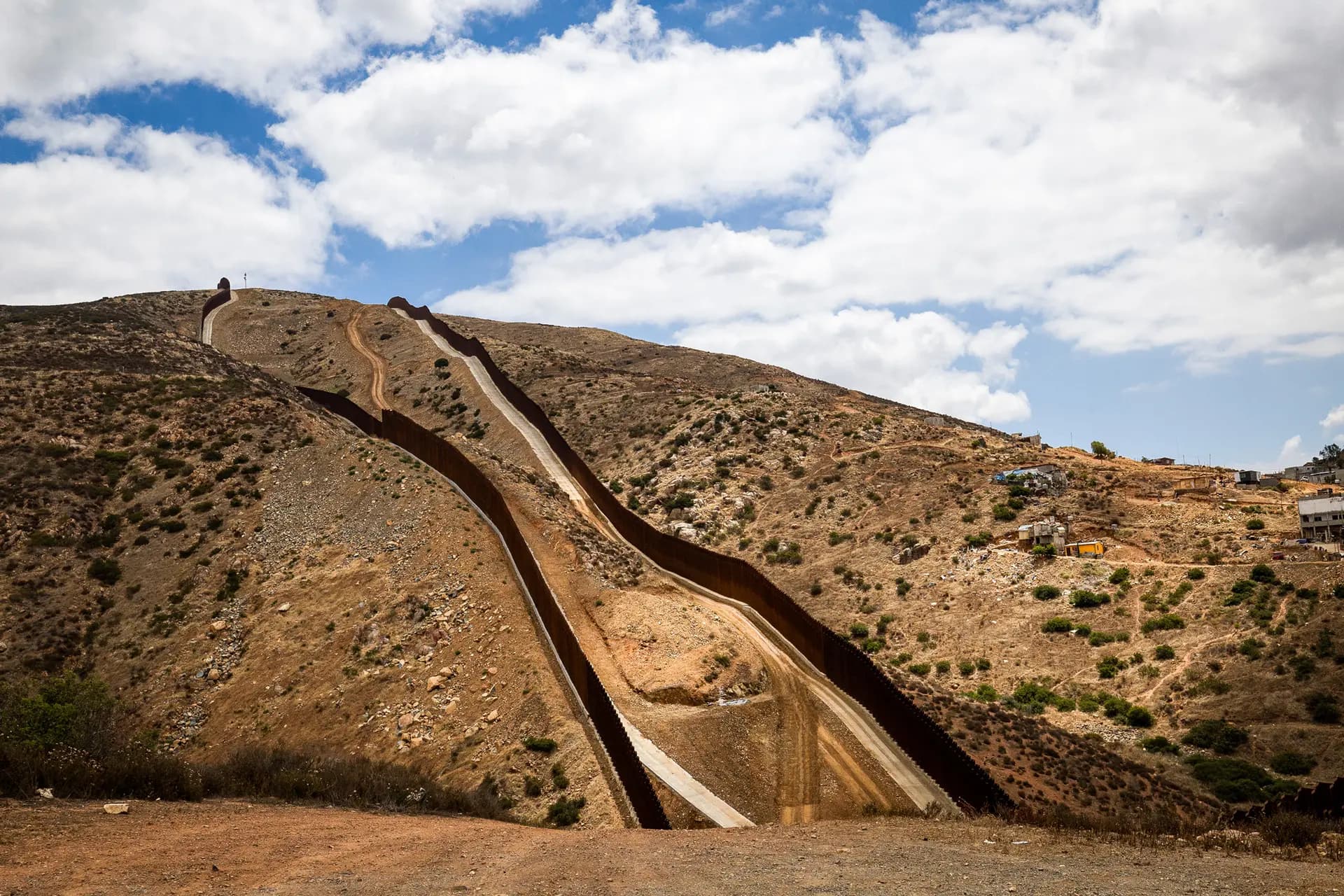 (Arriba) Vehículos de construcción circulan por un terreno montañoso a lo largo del muro fronterizo sur en las afueras de San Diego el 27 de mayo de 2026. (Abajo) El muro fronterizo sur, visto desde las afueras de San Diego el 27 de mayo de 2026. (John Fredricks/The Epoch Times)