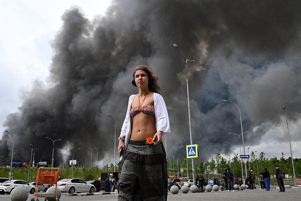 Una mujer camina por las inmediaciones de un centro comercial mientras se eleva humo negro desde la zona de la refinería de la petrolera rusa Gazprom Neft, situada en las afueras del sureste de Moscú, el 18 de junio de 2026. (AFP vía Getty Images).