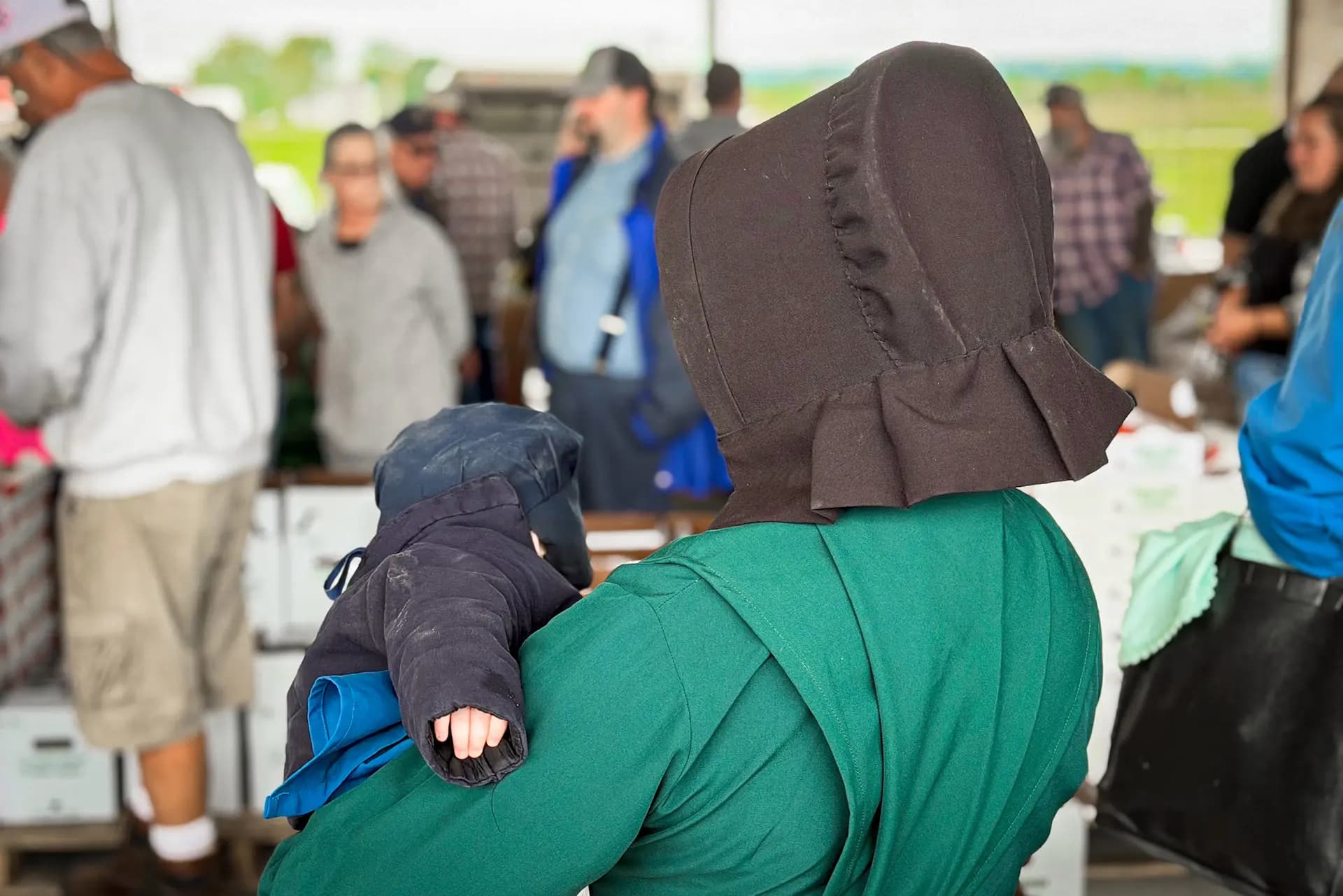 Una madre amish vestida con ropa tradicional sostiene a su hijo durante la subasta de productos agrícolas del valle de Mohawk el 16 de septiembre de 2026. (Allan Stein/The Epoch Times).