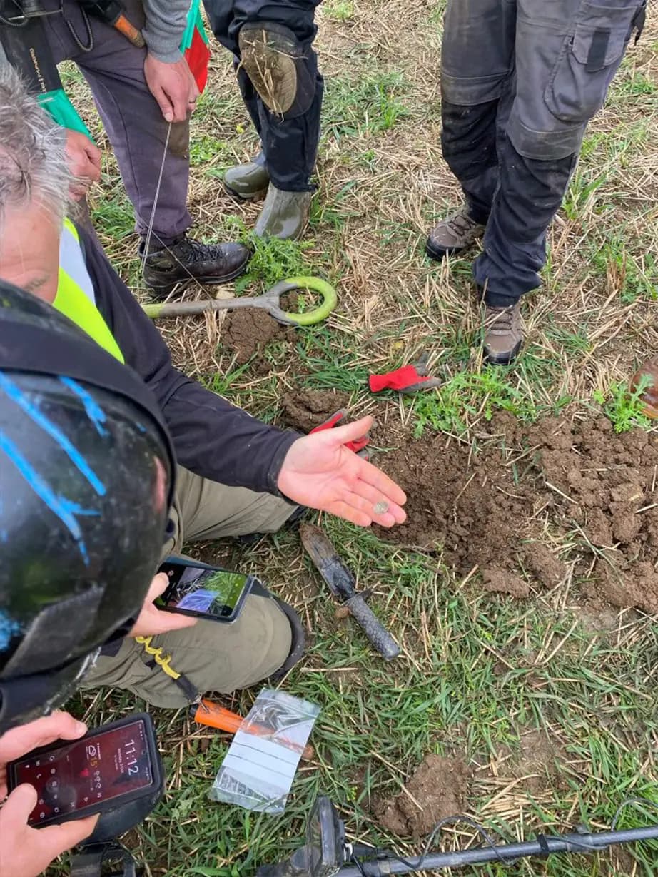 Unos aficionados a la detección de metales examinan una moneda de plata encontrada en un campo en Bickmarsh, Reino Unido. (Servicio de Archivo y Arqueología de Worcestershire)