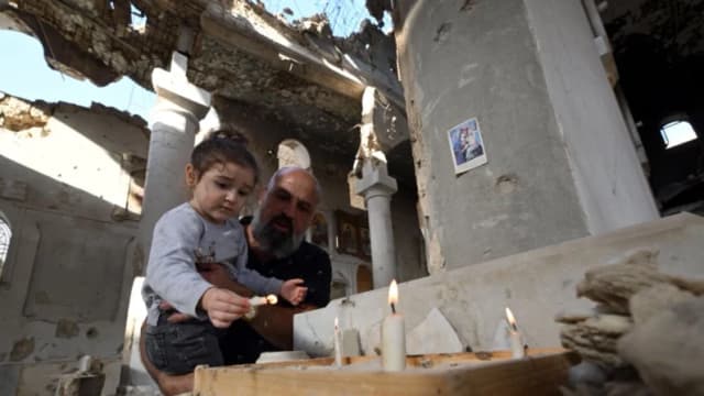Un cristiano sostiene a su hija mientras encienden velas en el altar de la iglesia ortodoxa griega de Saint Takla, en Darayya, a las afueras de Damasco, el 28 de octubre de 2025, mientras Siria se enfrenta a uno de sus retos más importantes: la reconstrucción bajo el nuevo liderazgo tras el derrocamiento del gobernante baazista Bashar al-Assad el pasado mes de diciembre. (Louai Beshara/AFP).