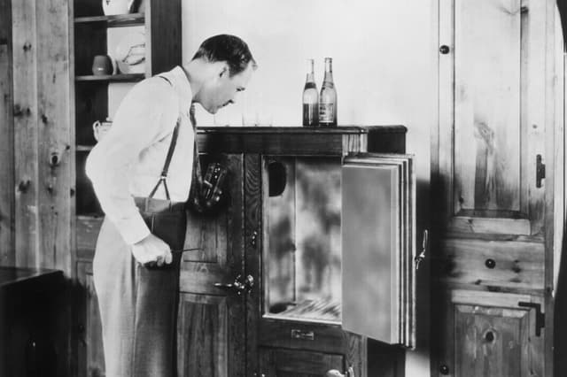 Un hombre con un vaso en la mano mira fijamente una nevera de madera vacía con dos botellas encima, alrededor de 1935. (Hulton Archive/Getty Images).