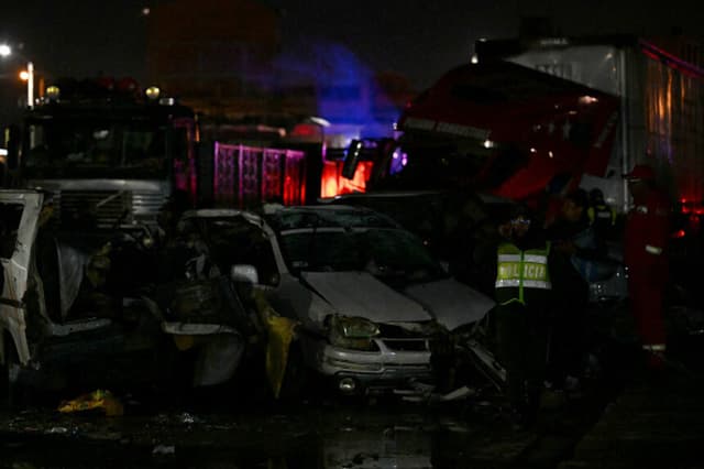 Un oficial de policía y equipos de rescate caminan junto a vehículos destrozados cerca del lugar donde se estrelló un avión militar en El Alto, cerca de La Paz, el 27 de febrero de 2026. Al menos 11 personas murieron el 27 de febrero cuando un avión de carga militar boliviano que transportaba billetes se estrelló al aterrizar cerca de la capital, La Paz. (AIZAR RALDES / AFP a través de Getty Images).
