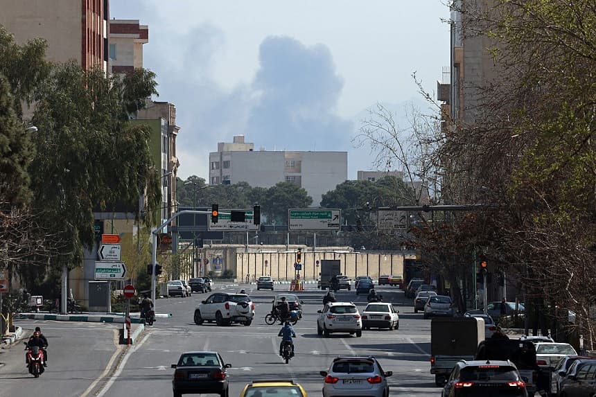 Los viajeros se abren paso por una calle concurrida con una nube de humo como telón de fondo tras los ataques aéreos en el centro de Teherán el 4 de marzo de 2026. (AFP vía Getty Images)