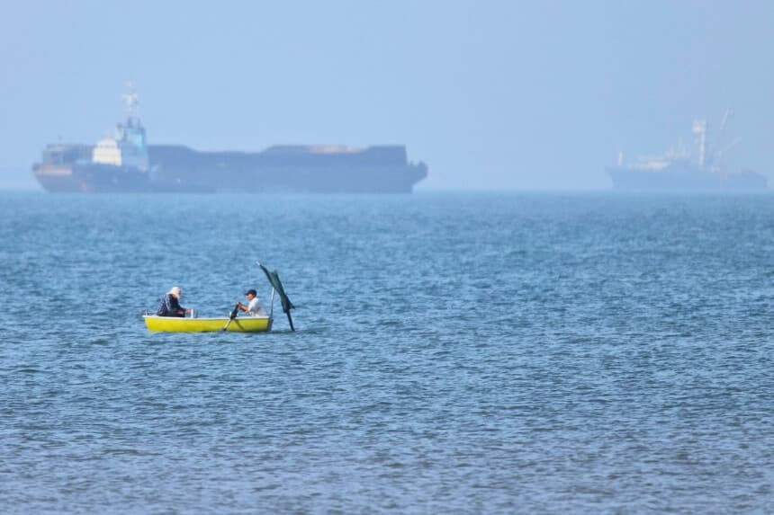 Dos hombres están sentados en una pequeña embarcación en el agua, con buques de carga fondeados al fondo, en el estrecho de Ormuz, frente a Bandar Abbas, Irán, el 4 de mayo de 2026. (Amirhosein Khorgooi/ISNA vía AP).