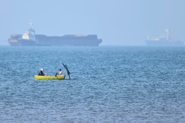 Dos hombres están sentados en una pequeña embarcación en el agua, con buques de carga fondeados al fondo, en el estrecho de Ormuz, frente a Bandar Abbas, Irán, el 4 de mayo de 2026. (Amirhosein Khorgooi/ISNA vía AP).