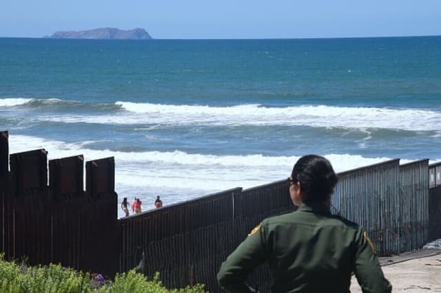 Imagen Ilustrativa: Un agente de la Oficina de Aduanas y Protección Fronteriza de EE. UU. vigila cerca de la estructura fronteriza que se adentra en el Océano Pacífico en San Diego, California, el 17 de abril de 2018, mientras personas visitan la playa en La Playa, México. La imagen fue tomada un día después de que California rechazara el despliegue de tropas de la Guardia Nacional en la frontera. (Frederic J. Brown / AFP vía Getty Images).