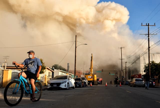 Una persona anda en bicicleta mientras un enorme incendio en un almacén de almacenamiento en frío sigue ardiendo y propagando humo por Los Ángeles el 20 de junio de 2026. (Mario Tama /Getty Images)