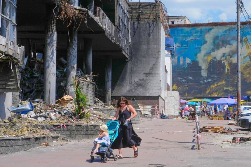 Una madre empuja un cochecito junto a un edificio dañado, cubierto de pinturas de artistas callejeros, y un gran mercado urbano que fue destruido recientemente por misiles rusos en Kiev, Ucrania, el 22 de junio de 2026. (Efrem Lukatsky/ AP Photo).