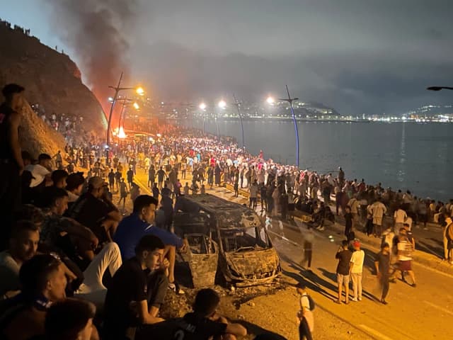 Los migrantes se reúnen alrededor de un vehículo incendiado mientras esperan la oportunidad de cruzar al enclave español de Ceuta, en el lado marroquí de la frontera en Fnideq, Marruecos, el 31 de julio de 2026. (Ahmed El Jechtimi /Reuters)