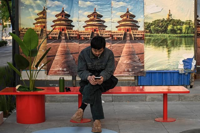 Un hombre revisa su teléfono móvil mientras descansa en un banco a lo largo de una calle en Pekín el 6 de mayo de 2025. (Jade Gao/AFP vía Getty Images).