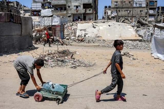 Unos niños transportan una carreta cargada con recipientes de agua junto a tiendas de campaña y escombros en un campamento para personas desplazadas por la guerra en el barrio de Sheikh Radwan, en la ciudad de Gaza, el 6 de julio de 2026. (Omar al-Qattaa/AFP vía Getty Images)