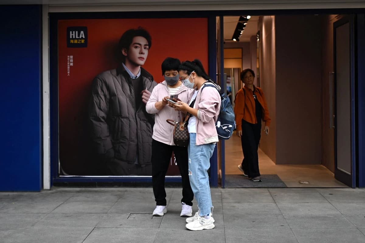 Mujeres usan sus teléfonos móviles frente a una tienda de ropa en una zona comercial de Beijing el 15 de septiembre de 2025. (Wang Zhao/AFP vía Getty Images).