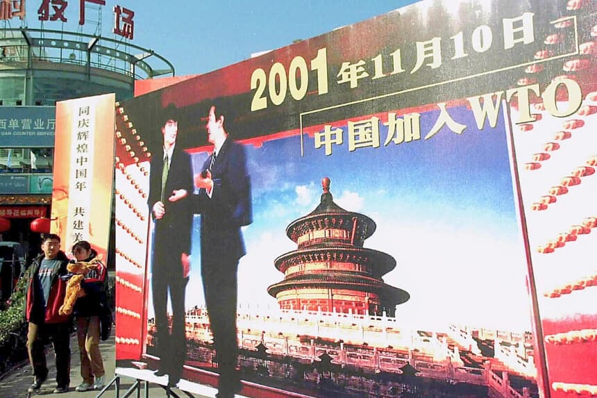 Varias personas pasan junto a una valla publicitaria que celebra la entrada de China en la Organización Mundial del Comercio en 2001 en Pekín, el 5 de enero de 2002. (AFP vía Getty Images).